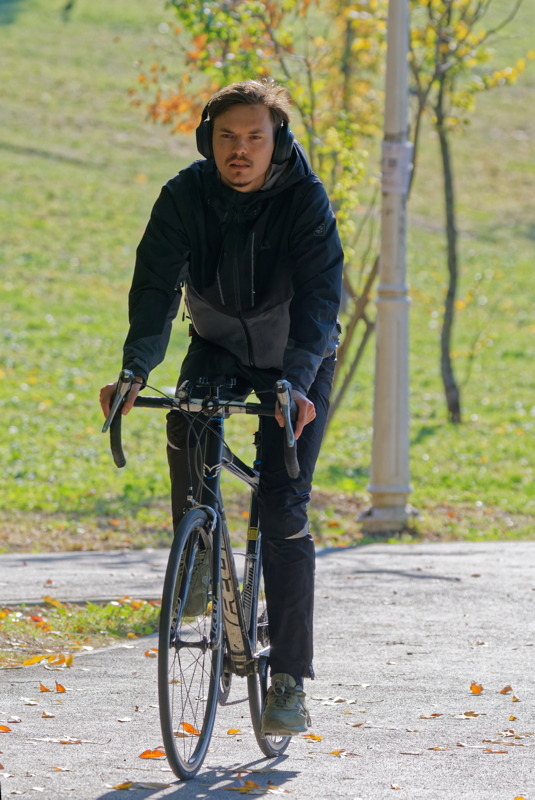 17 - A man riding a road bike on a paved path outdoors while wearing black outerwear and headphones